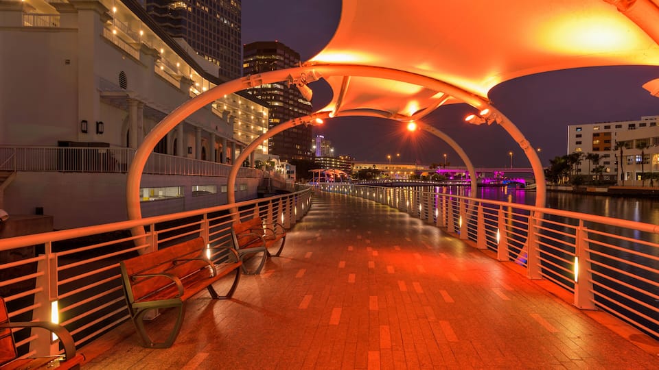 A nighttime waterfront promenade with illuminated arches, benches, and a river.