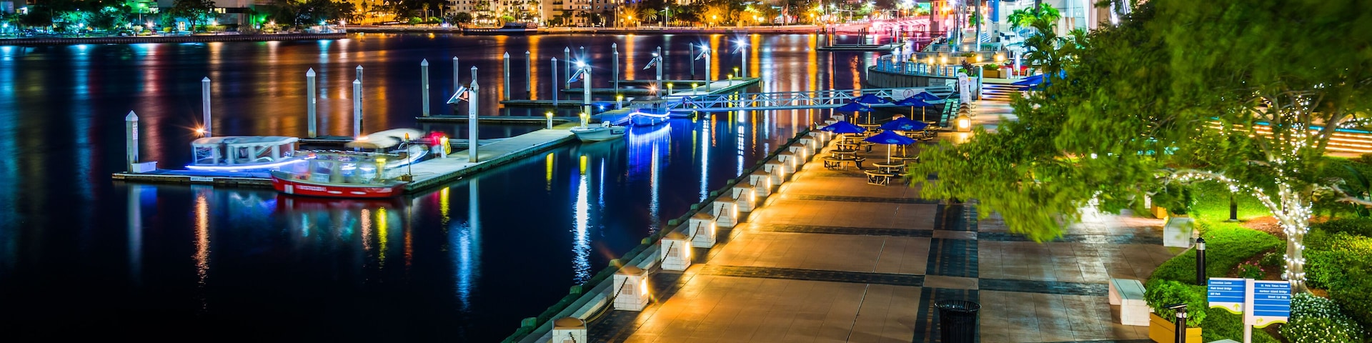 Um calçadão à beira-mar à noite, com edifícios iluminados, um cais com barcos e vista clara para a água.