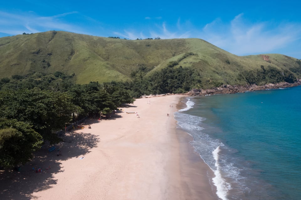 A beach with a sandy shore, a forested hill, and a clear blue ocean.