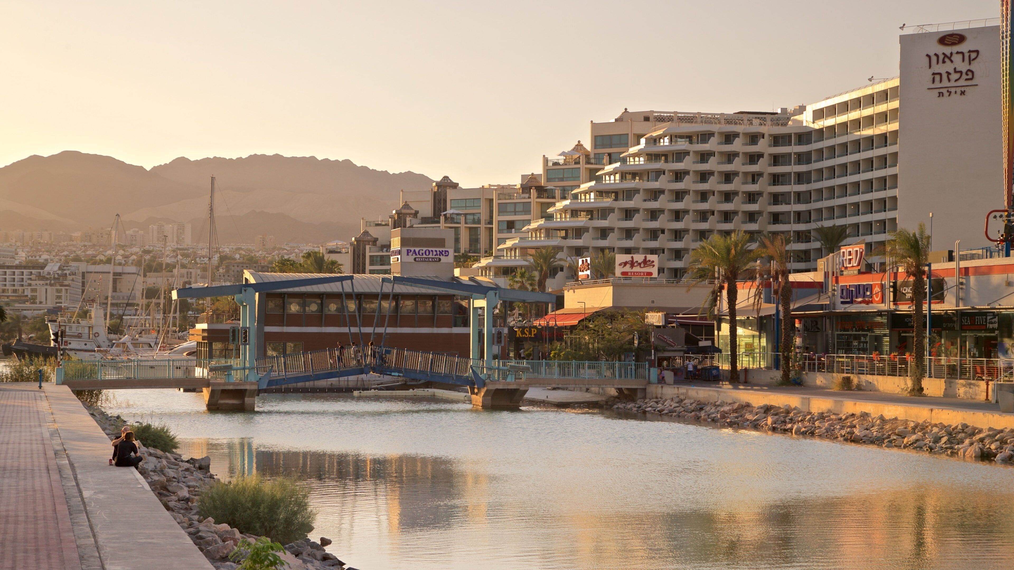 Un paseo ribereño con un puente peatonal, un restaurante y un hotel.