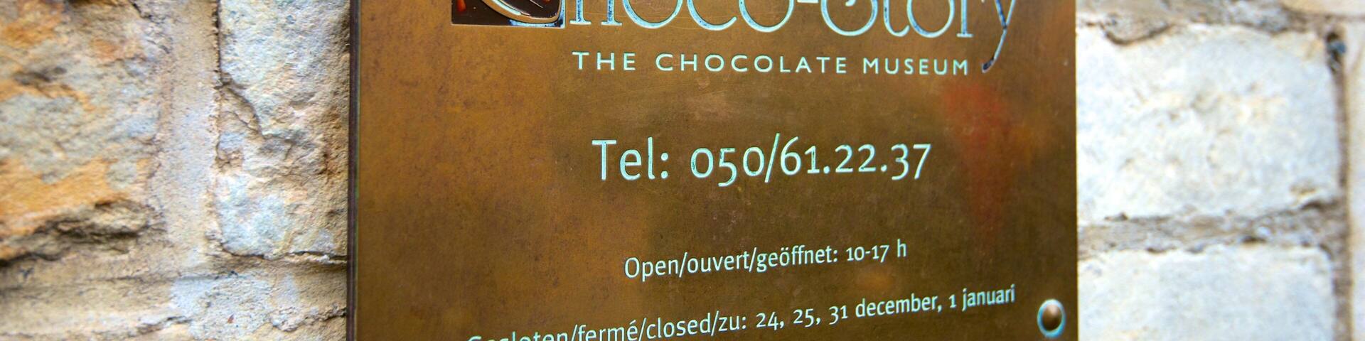 A plaque on a stone wall for Choco-Story, the Chocolate Museum, with contact information and opening hours.