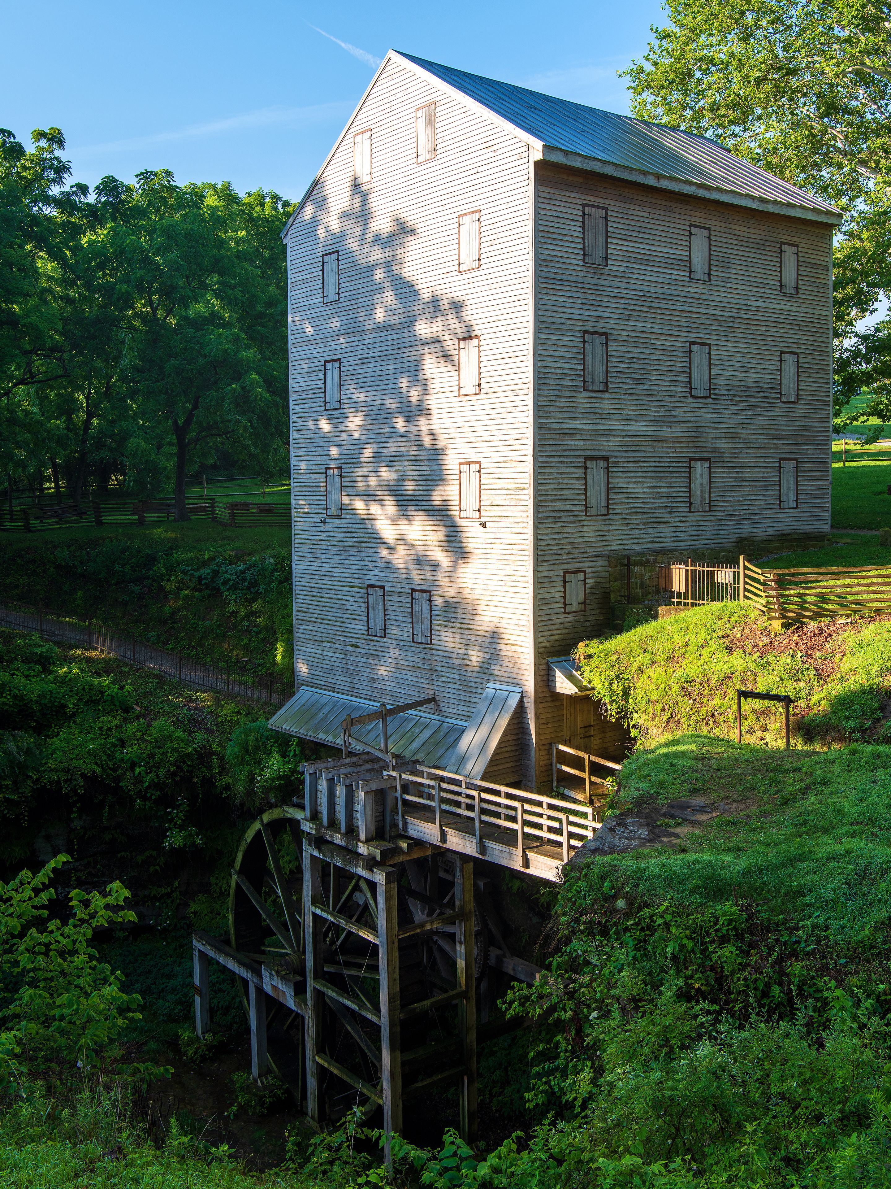 A wooden watermill with a wooden staircase leading up to it, surrounded by lush greenery and trees.