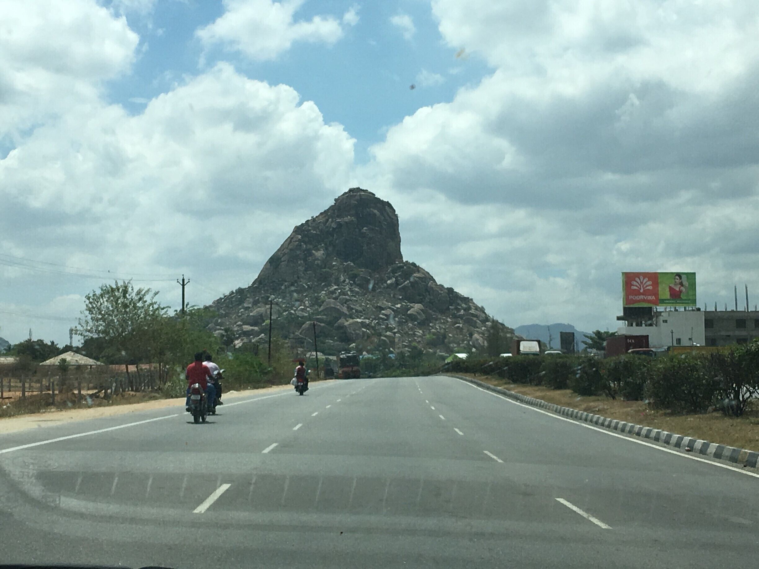 A road with a large rock formation in the distance, a billboard, and a clear sky.