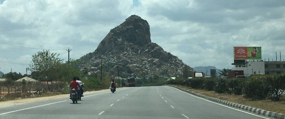 A road with a large rock formation in the distance, a billboard, and a clear sky.