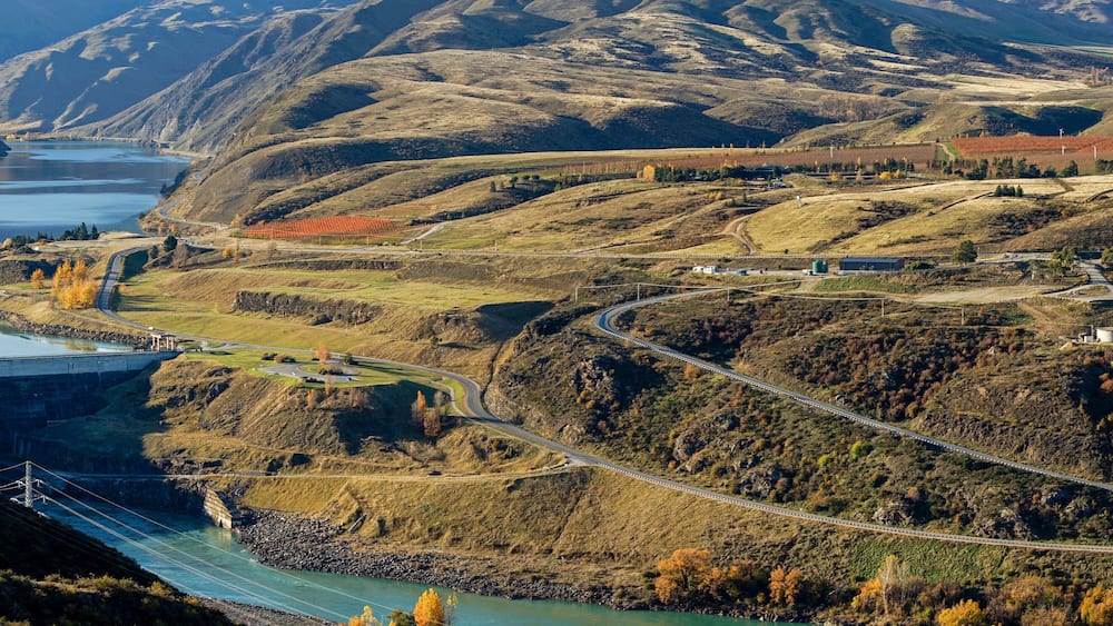 Panoramic aerial view to Clyde dam with river and winding road on hillside surrounded by mountainous and autumn trees.