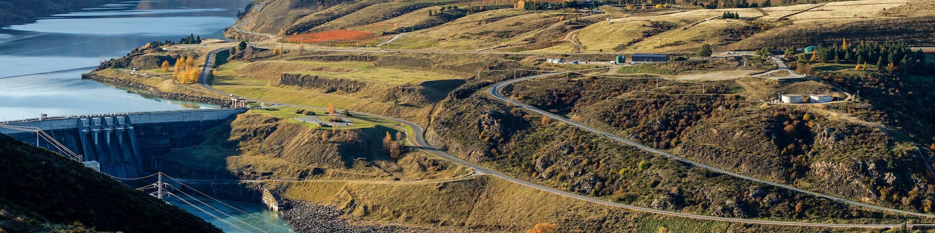 Panoramic aerial view to Clyde dam with river and winding road on hillside surrounded by mountainous and autumn trees.