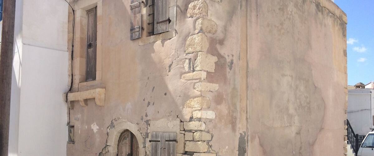 A weathered building with stone walls, wooden shutters, and a single arched door.