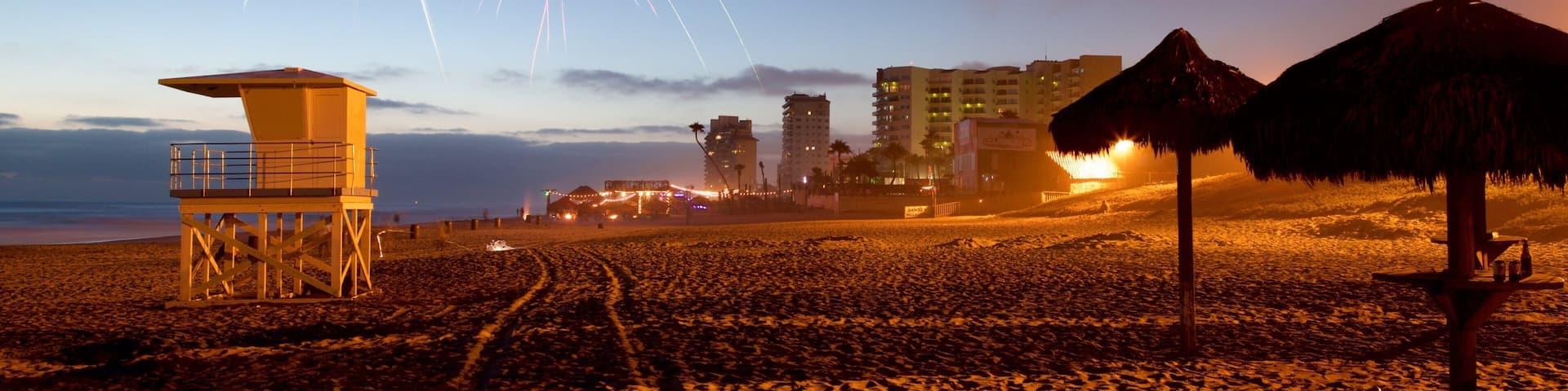 A beach with a lifeguard tower, a thatched umbrella, and fireworks in the sky.