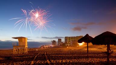 Una playa con una torre de salvavidas, una sombrilla de paja y fuegos artificiales en el cielo.