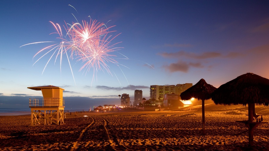 Una spiaggia con una torretta di salvataggio, un ombrellone di paglia e fuochi d'artificio nel cielo.