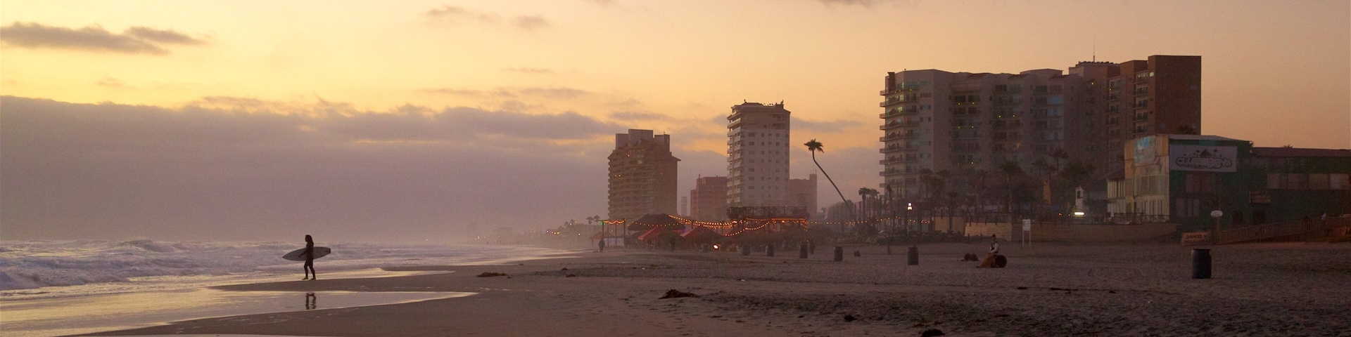 A beach at sunset with a person carrying a surfboard, buildings in the background, and palm trees.