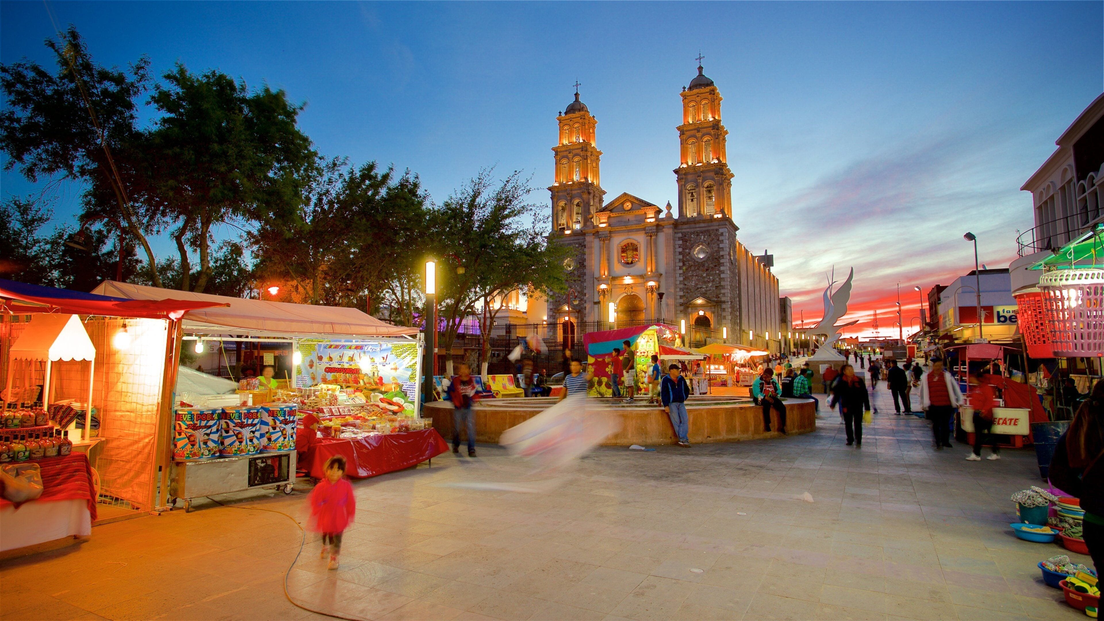 A bustling evening market with stalls and a historic church with two towers in the background.