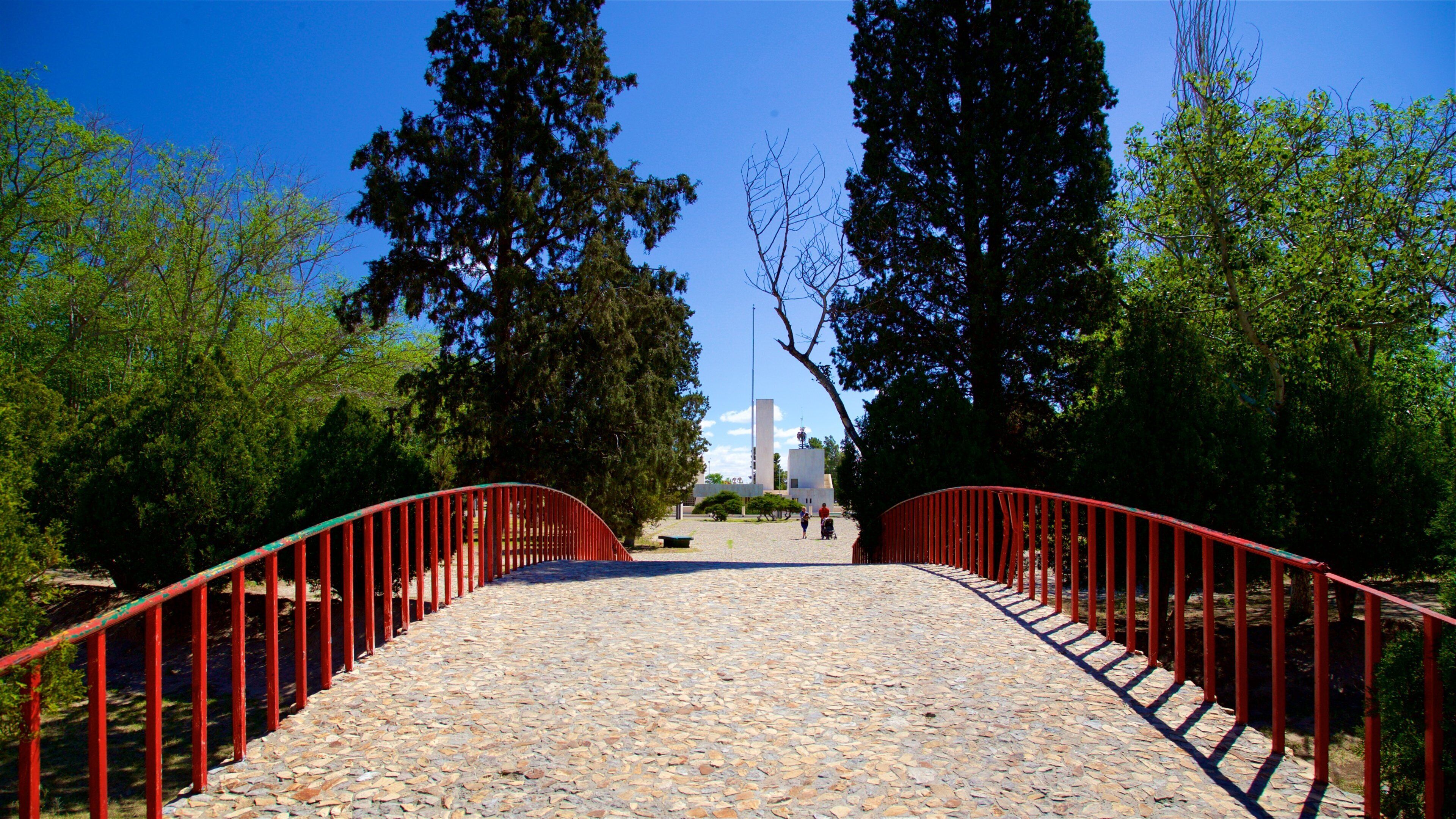 Un sendero adoquinado con barandillas rojas, rodeado de árboles y que conduce a un edificio con torre.