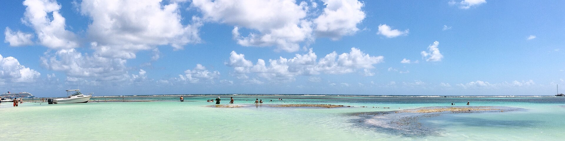 Una playa con agua turquesa cristalina, algunas personas y barcos a lo lejos.