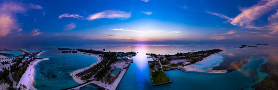 Un resort tropical con aguas turquesas cristalinas, un muelle largo y una puesta de sol sobre el océano.
