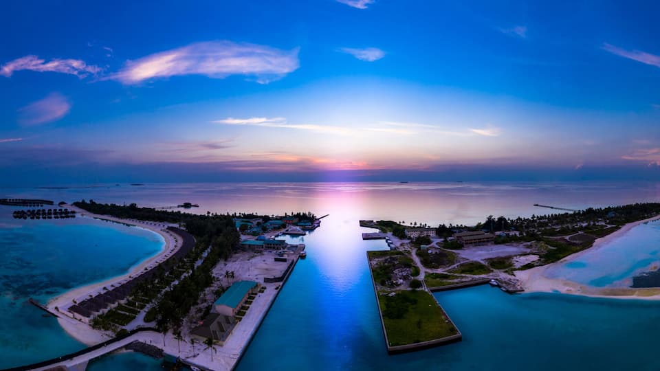 A tropical resort with clear turquoise waters, a long pier, and a sunset over the ocean.