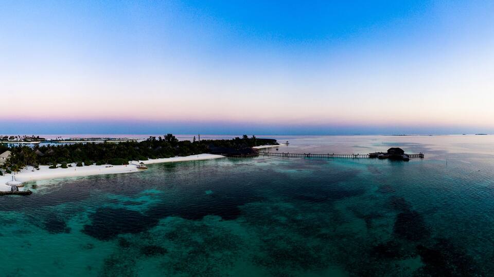 An aerial view of a tropical resort with overwater bungalows, a white sandy beach, and clear turquoise waters.
