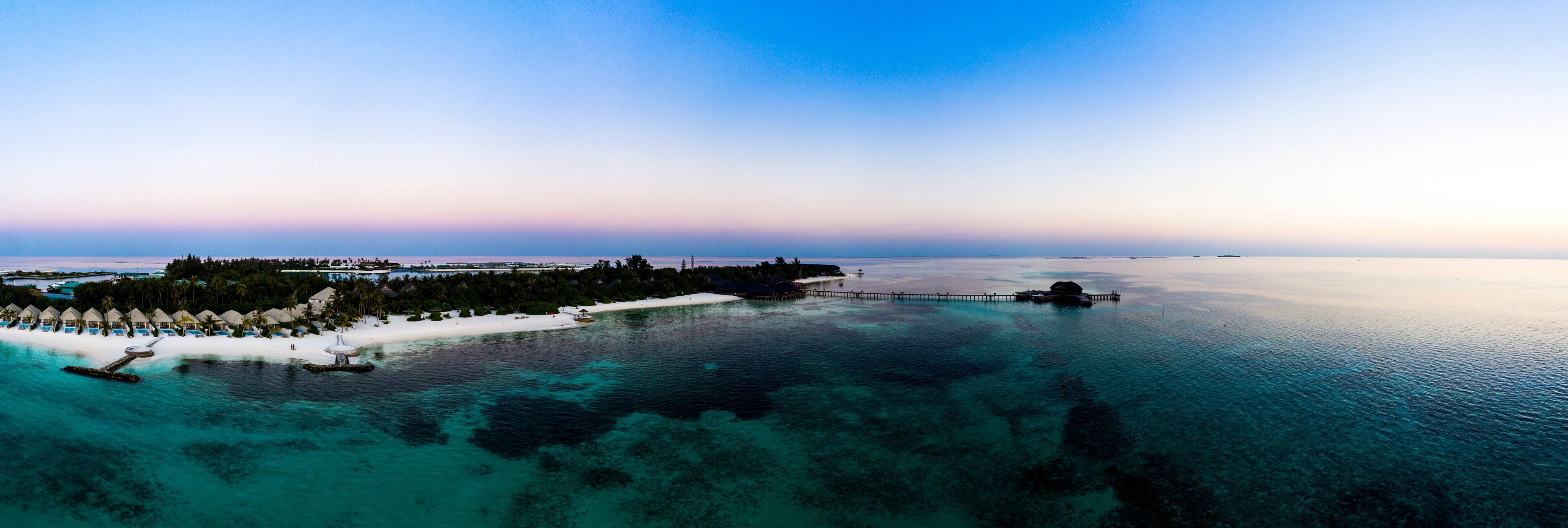 An aerial view of a tropical resort with overwater bungalows, a white sandy beach, and clear turquoise waters.