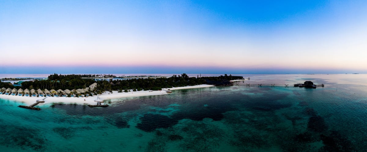 An aerial view of a tropical resort with overwater bungalows, a white sandy beach, and clear turquoise waters.