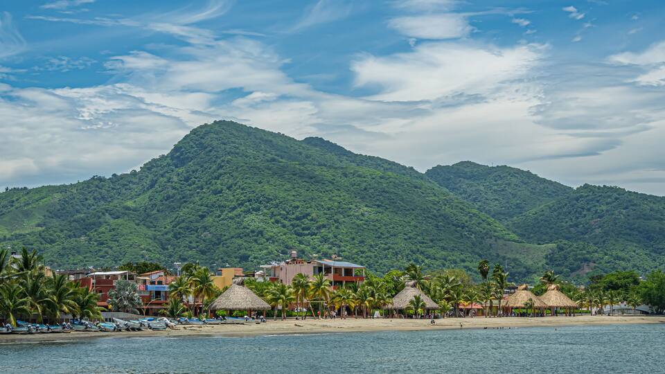 Zihuatanejo, Mexico - July 18, 2023: Playa La Madera with straw covered circular open-wall huts to offer shade in front of green forested hills under blue cloudscape. Boats and houses