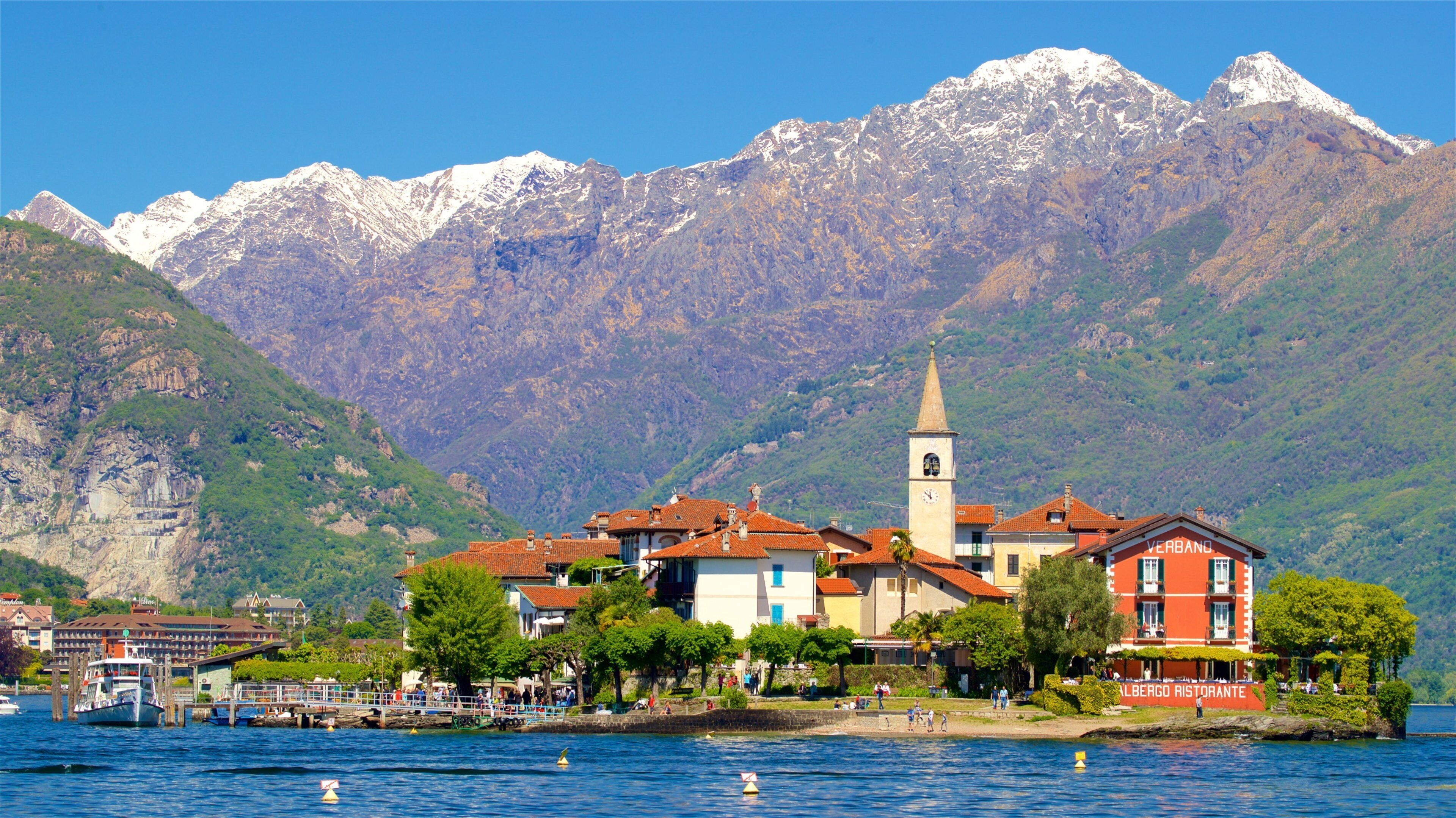 A lakeside town with a prominent church tower, a restaurant named "Albergro Ristorante," and snow-capped mountains in the background.