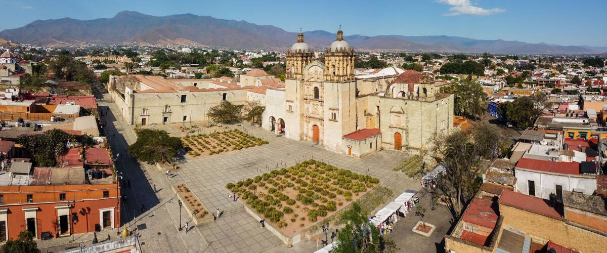 A historic church with two domes, a plaza with a garden, and mountains in the background.