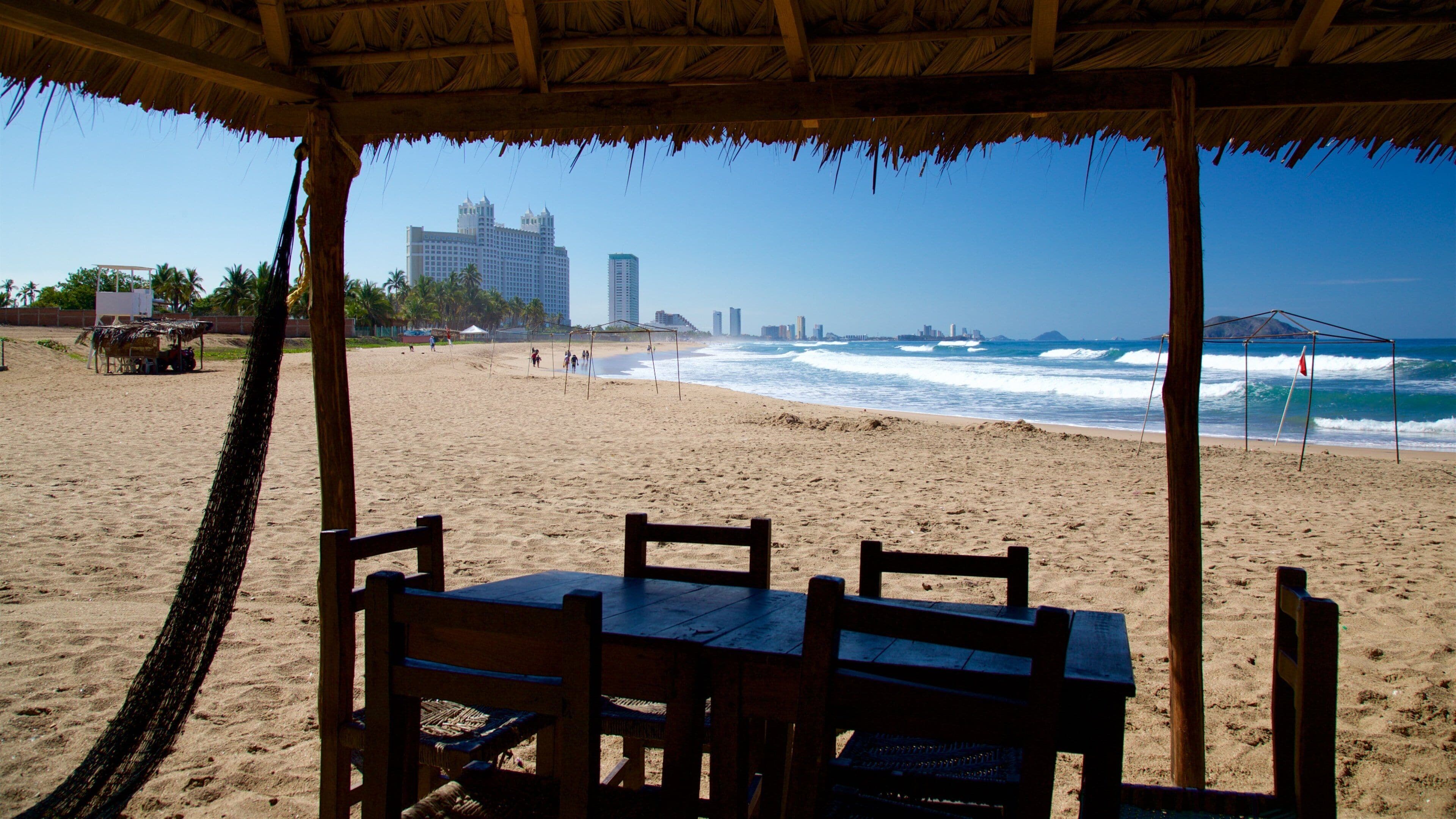 A beach with a wooden table and chairs under a thatched roof, overlooking the ocean with waves and a city skyline in the background.