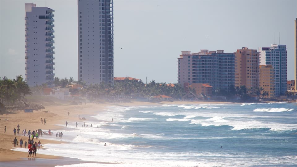 A beach with waves, palm trees, and tall buildings in the background.
