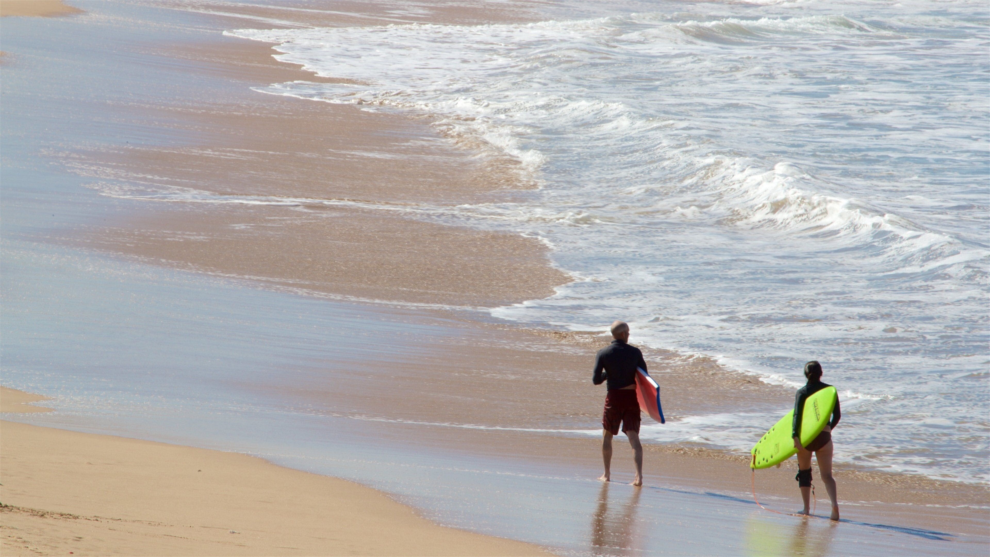 A beach with waves and two people carrying surfboards.