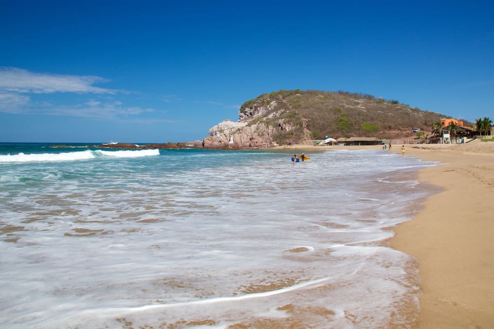 A beach with waves, a rocky hill, and palm trees.