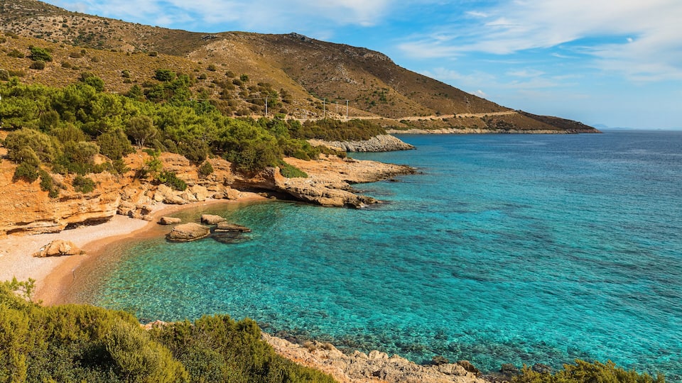 A coastal landscape with clear turquoise waters, rocky beach, and green vegetation on a hillside.