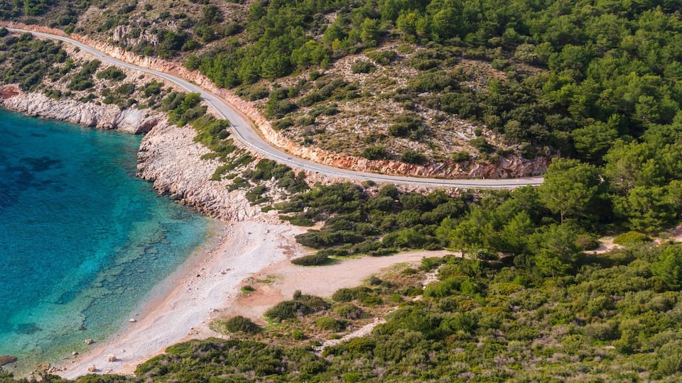 A coastal road winding through a forested area with a beach and turquoise waters.