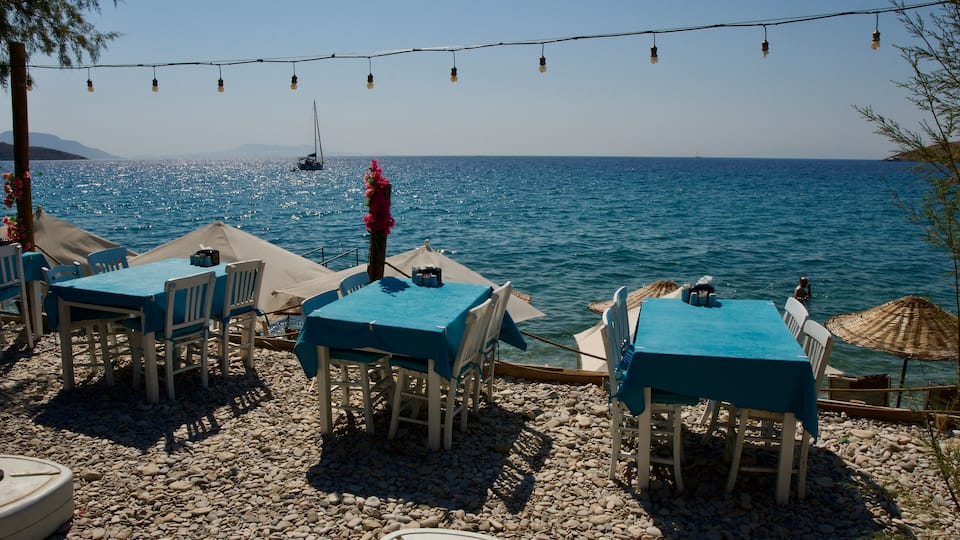 A seaside restaurant with tables and chairs, a boat in the background, and a clear blue sky.