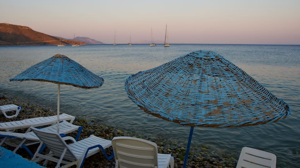 A beach with lounge chairs and blue woven umbrellas, a calm sea, and sailboats in the distance.