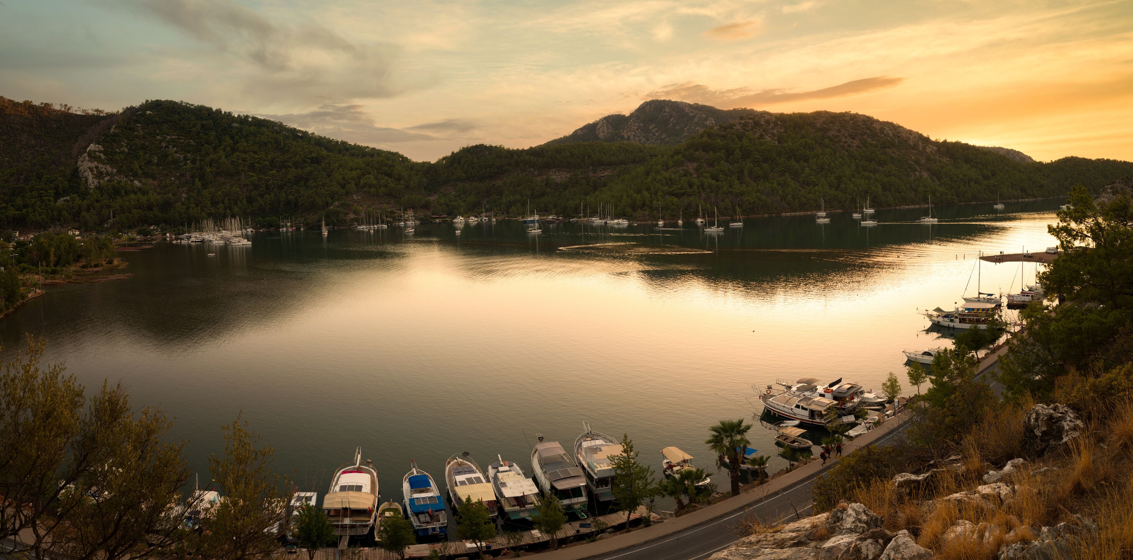 A calm lake with boats docked along the shore, surrounded by hills and trees.