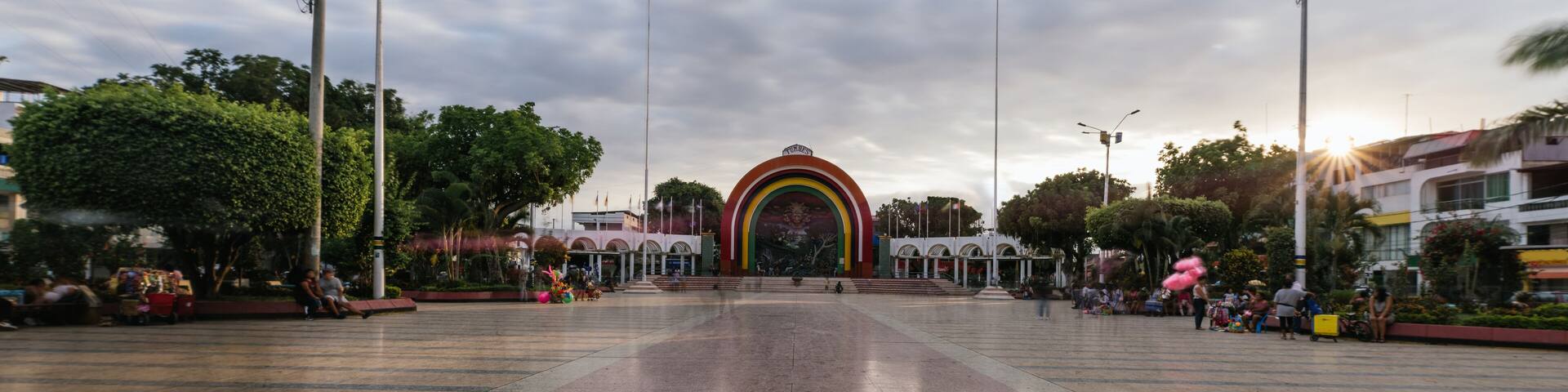 Una plaza con un arco colorido, árboles y farolas.