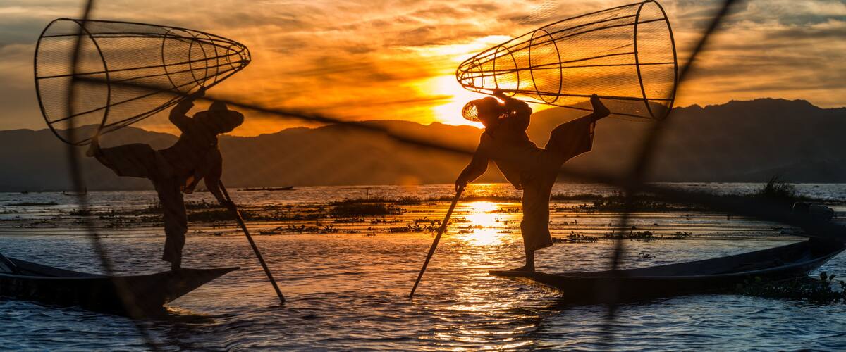 Two people on a boat at sunset with fishing nets overhead.