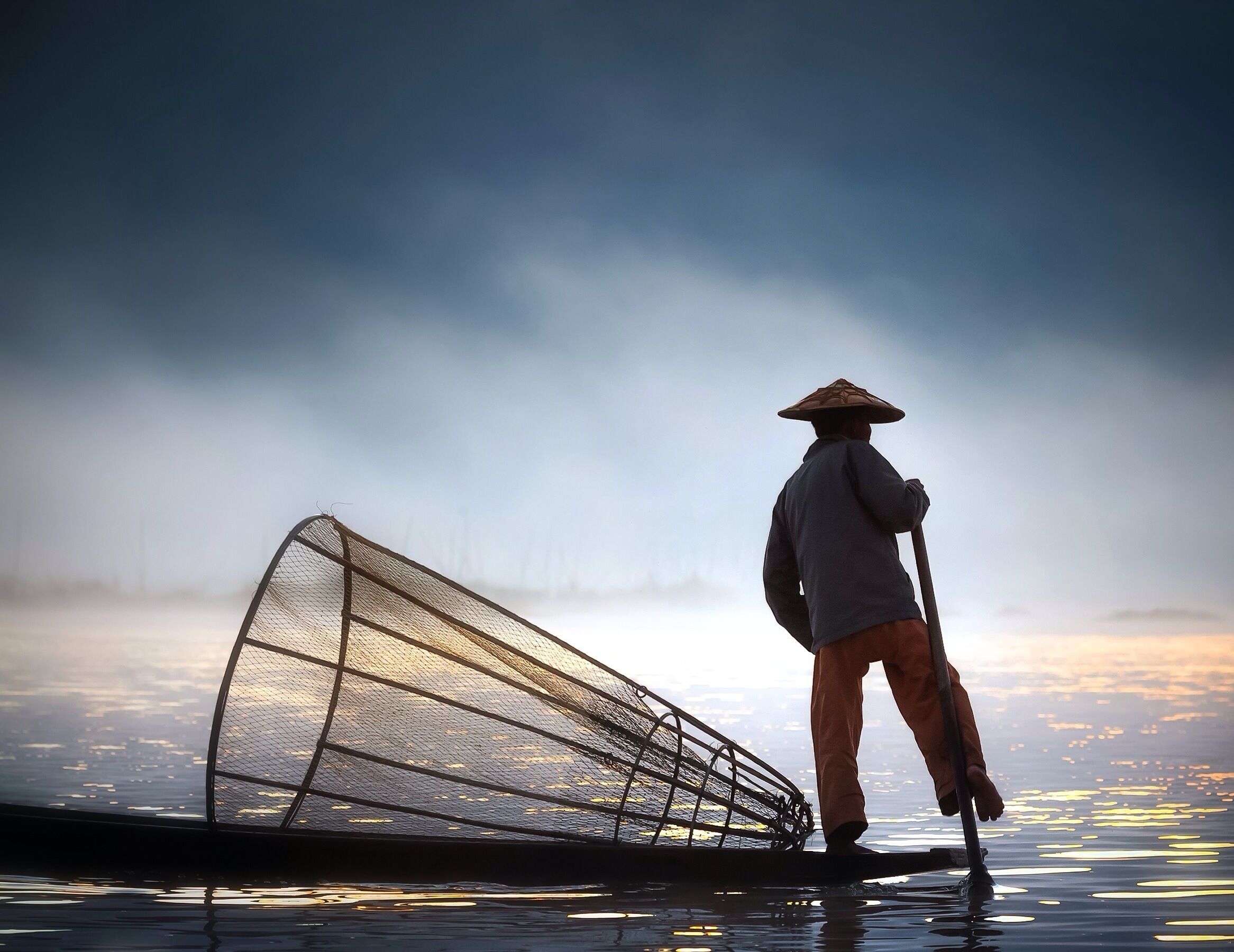 A person standing on a boat with a fishing net, wearing a conical hat, in a body of water with a misty background.