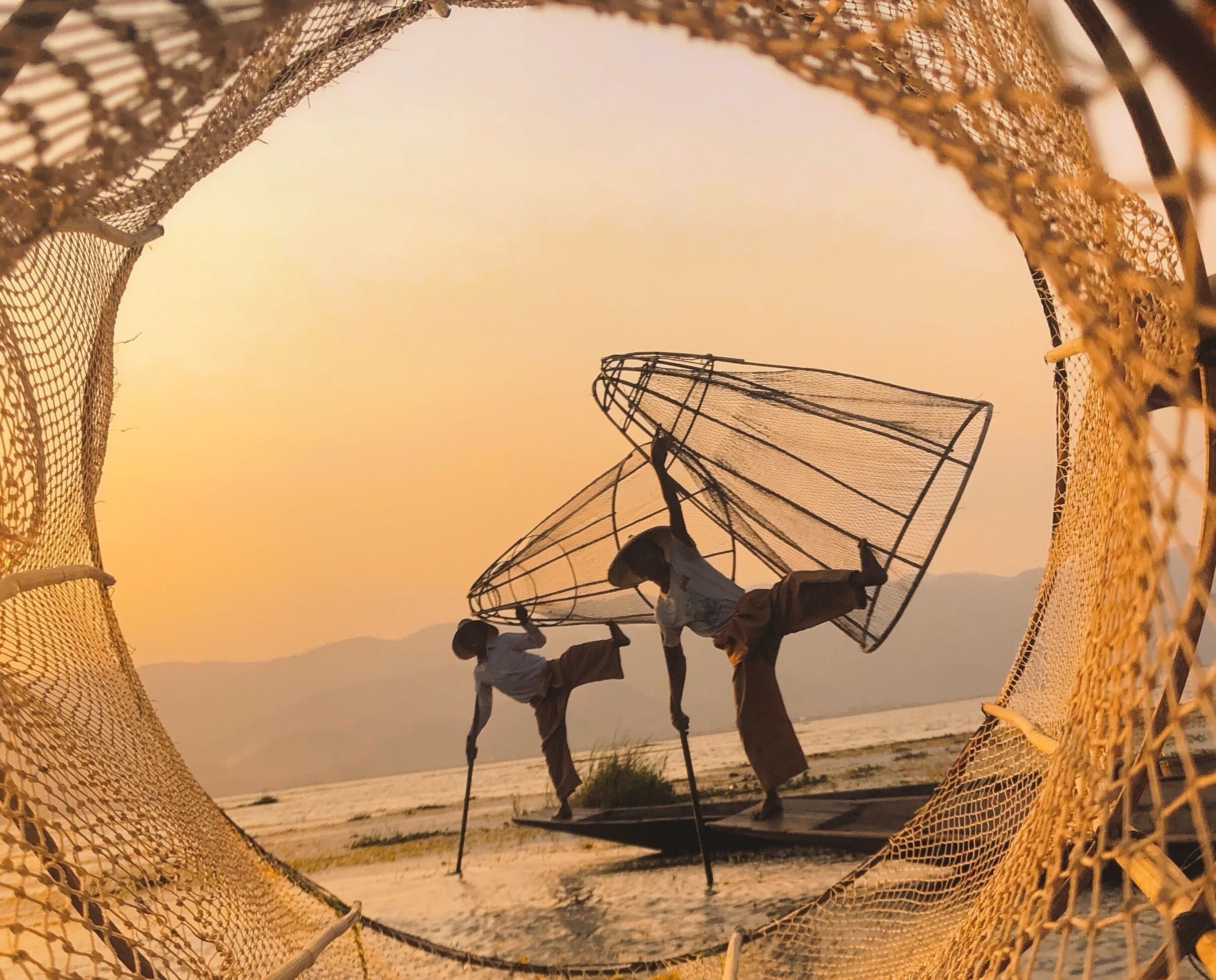 Two people carrying fishing nets on a beach with mountains in the background.