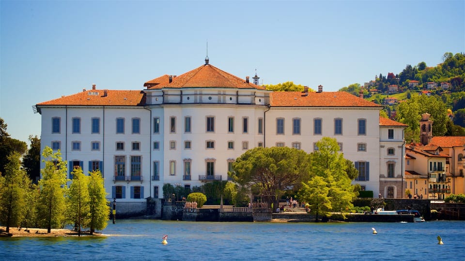 A large white building with a red roof by a calm lake, surrounded by trees and other buildings.