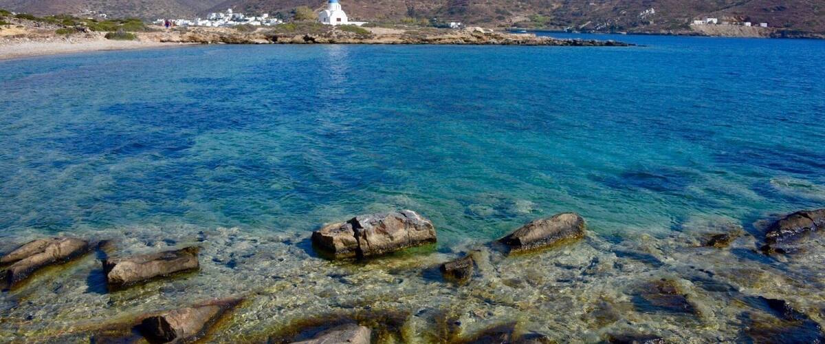 A coastal scene with clear blue water, rocky shoreline, and a white building with a blue dome on a hill.