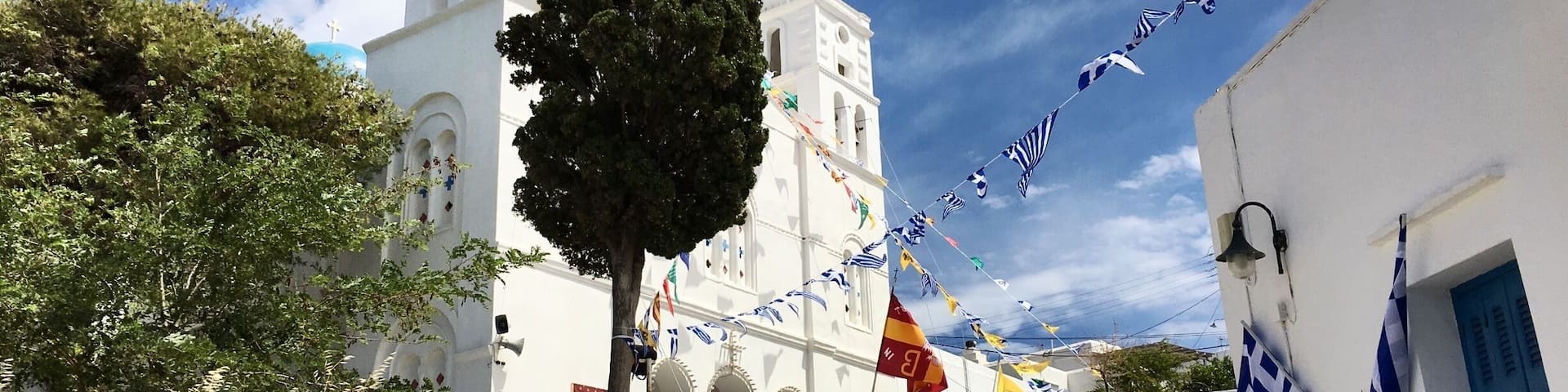 A white church with a clock tower, a stone wall, and a tree. Flags are strung across the street.