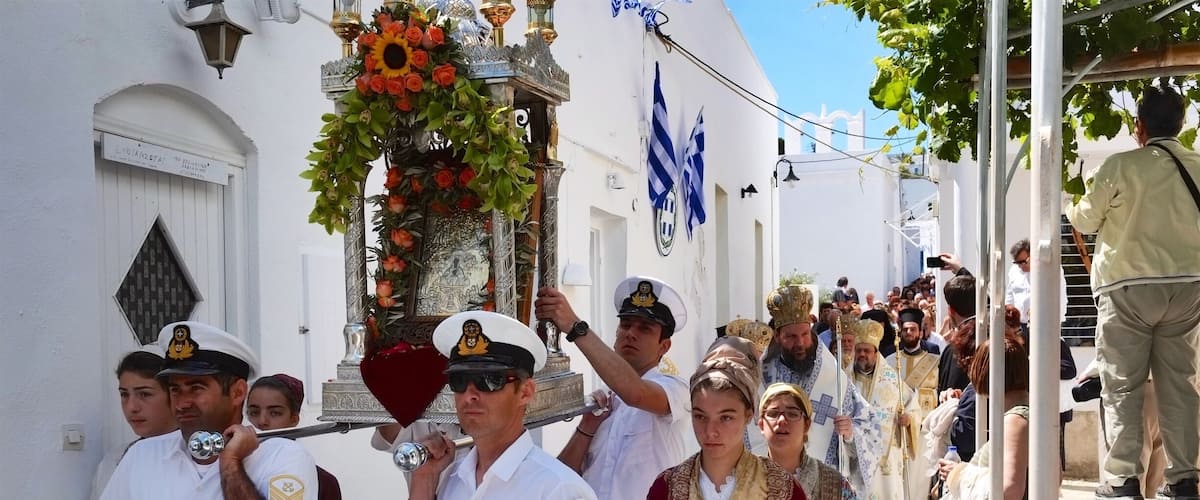 A street parade with participants in traditional attire, a white building with blue and white flags, and a crowd of onlookers.
