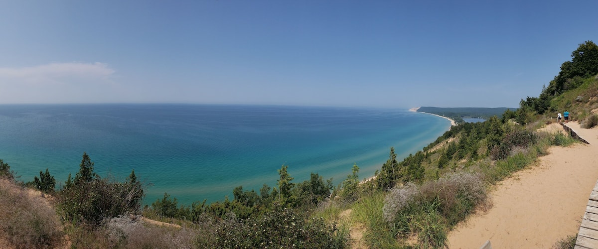 A coastal landscape with a sandy path, dense greenery, and a clear view of the ocean.