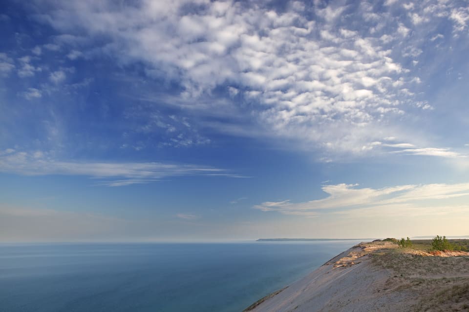 Un paisaje costero con una playa arenosa, dunas cubiertas de hierba y un cielo azul despejado con algunas nubes.