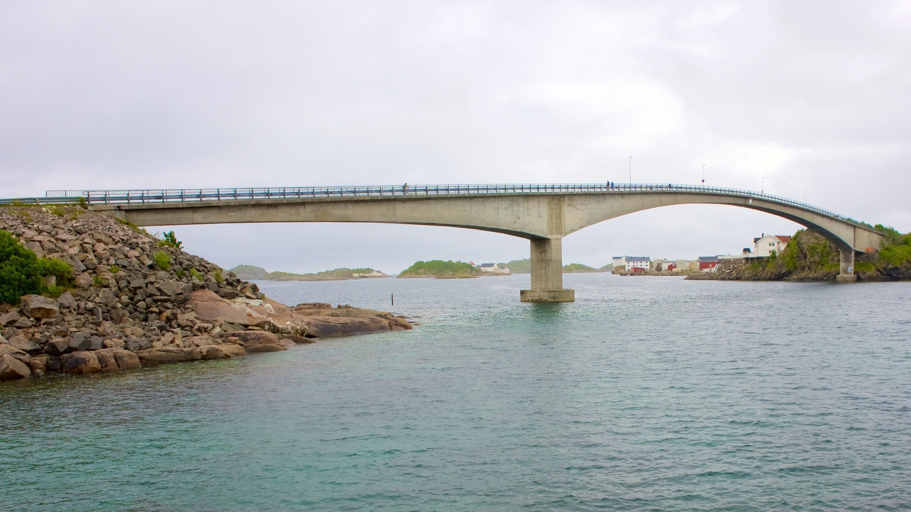 A concrete bridge over a calm river with rocky shores and distant buildings.