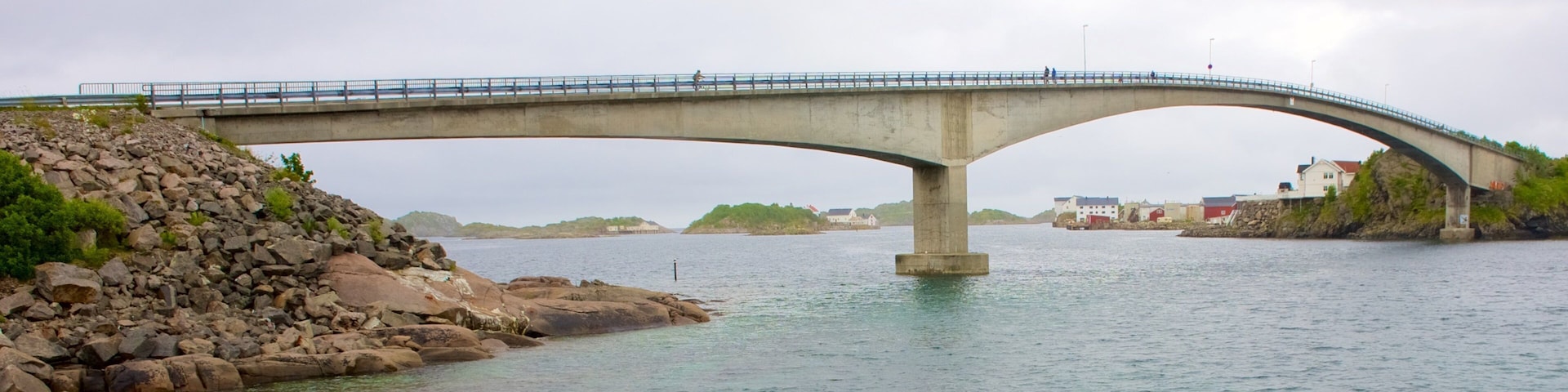 A concrete bridge over a calm river with rocky shores and distant buildings.