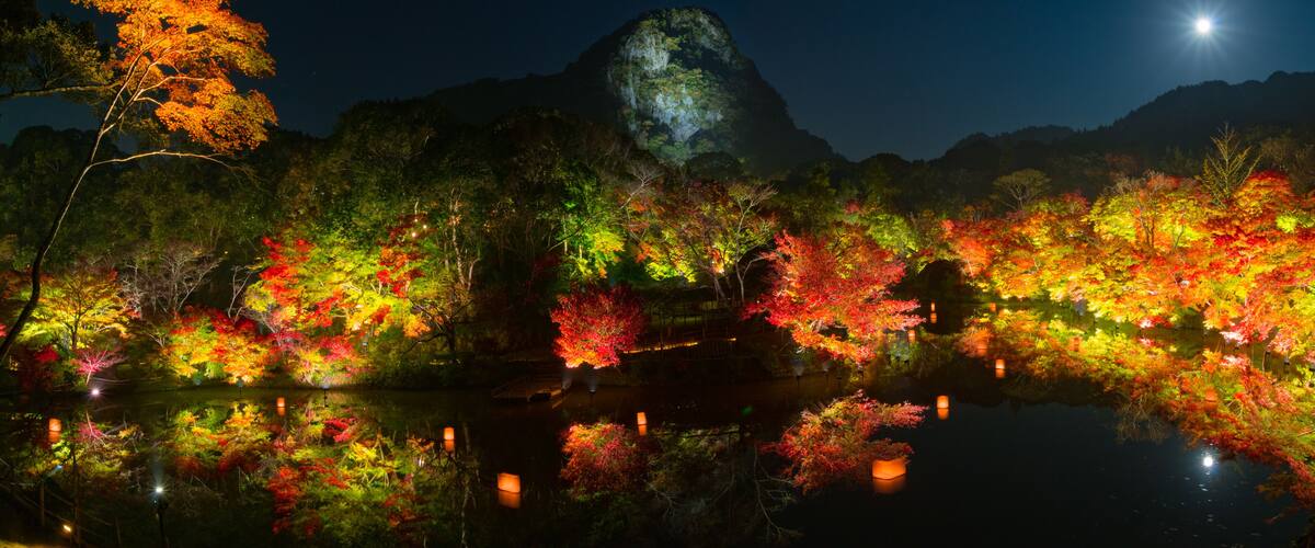 A night scene with a mountain, colorful autumn trees, and a calm lake reflecting the lights.