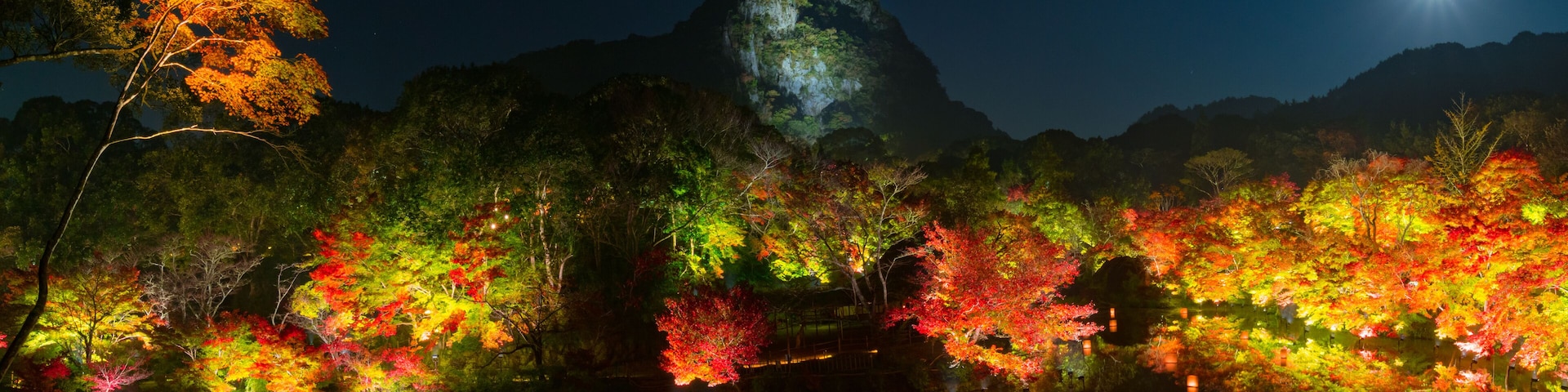 A night scene with a mountain, colorful autumn trees, and a calm lake reflecting the lights.