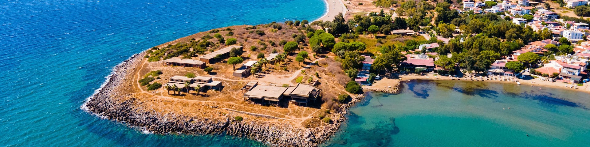 An aerial view of a coastal town with a prominent beach, a small island with buildings, and a mountainous backdrop.
